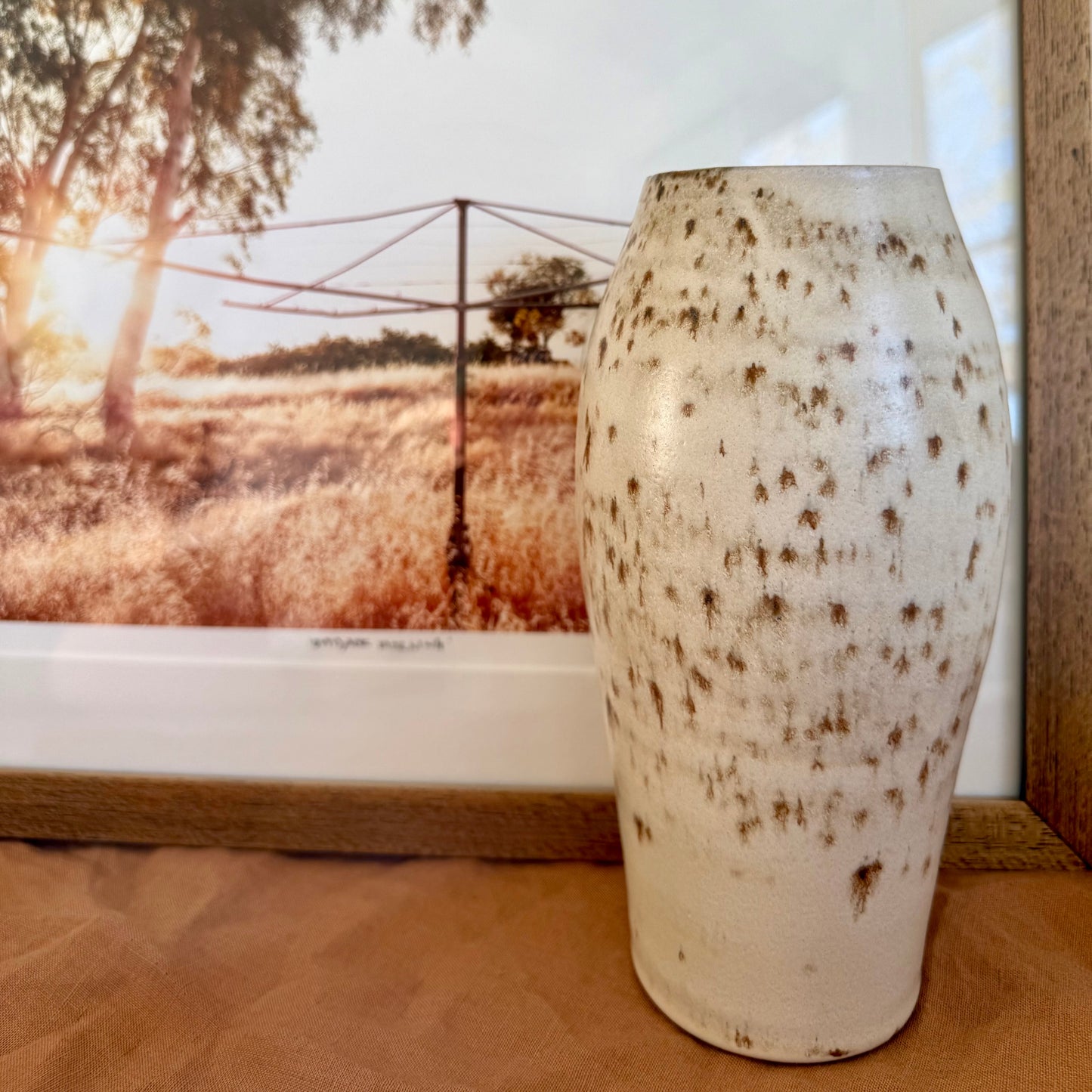 White ceramic vase with brown speckles on a wooden surface, with a scenic window view in the background.