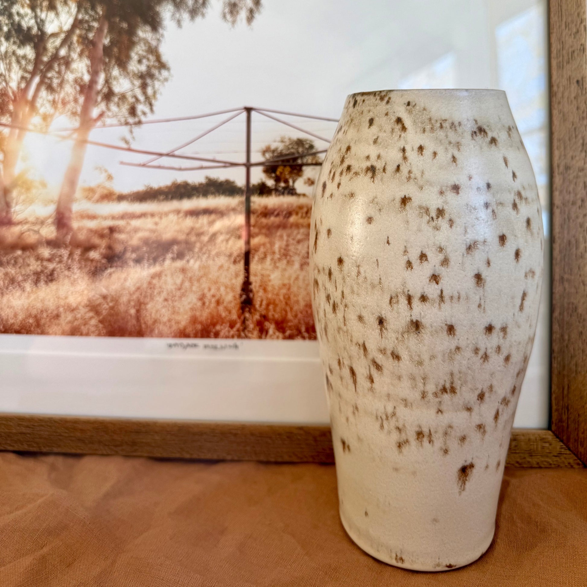 White ceramic vase with brown speckles on a wooden surface, with a scenic window view in the background.